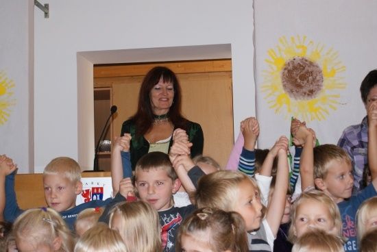 Eine Frau steht hinter einem Rednerpult mit Mikrofon und lächelt eine Gruppe von Kindern an, die ihre Hände in der Luft heben. Die Kinder befinden sich in einem Klassenzimmer mit einem Sonnenblumenbild an der Wand.