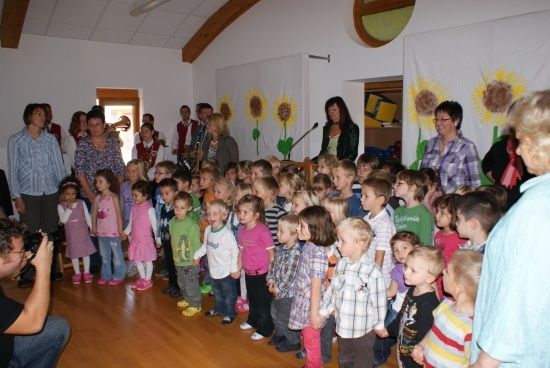 Eine Gruppe von Kindern und Erwachsenen versammelt sich in einem Raum mit Holzböden, Sonnenblumen an der Wand und einer Bühne mit Mikrofon und Trompete.