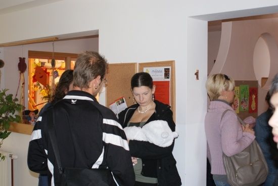 Eine Gruppe von Menschen steht in einem Raum, möglicherweise bei einer Veranstaltung. Eine Frau schaut auf etwas an der Wand, während ein Mann sie ansieht. Eine andere Frau hält ein Baby. An der Wand hängen Poster.