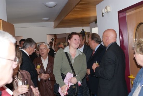 Eine Gruppe von Menschen steht und unterhält sich in einem Raum, einige halten Gläser mit Getränken. Eine Frau in der Mitte lächelt und hält ein rosa Tuch.