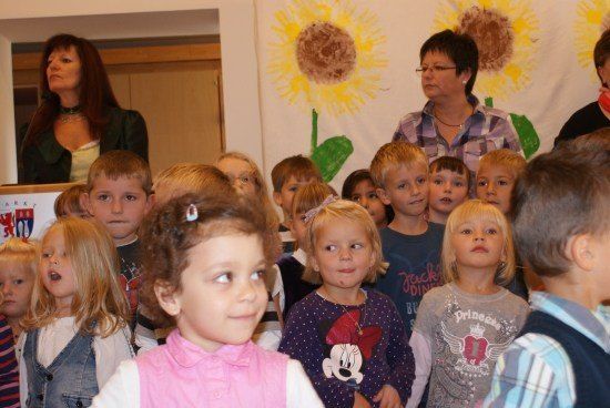 Eine Gruppe von Kindern und zwei Frauen steht in einem Raum mit einem Sonnenblumenhintergrund. Die Kinder schauen nach oben, möglicherweise singen oder lauschen sie.