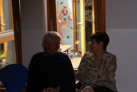 Zwei Frauen sitzen in einem Raum mit einem Fenster im Hintergrund. Die Frau auf der linken Seite trägt einen schwarzen Schal und die Frau auf der rechten Seite trägt eine Brille.