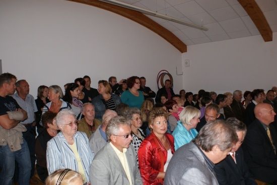 Eine große Gruppe von Menschen ist in einem Raum versammelt, einige stehen und einige sitzen, wobei eine Frau in einer roten Jacke vorne heraussticht.
