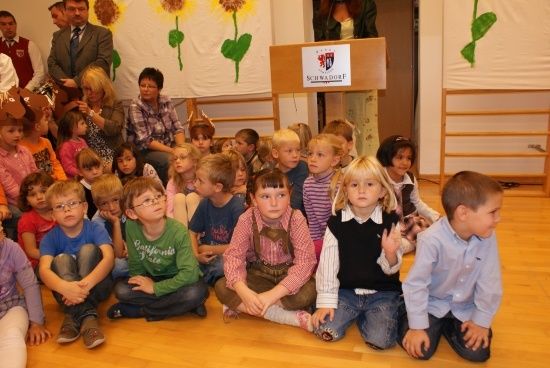 Eine Gruppe von Kindern in einem Klassenzimmer, einige mit Brille, sitzen auf dem Boden. Ein Podium mit einem Schild, das SCHWADORF liest, ist im Hintergrund.