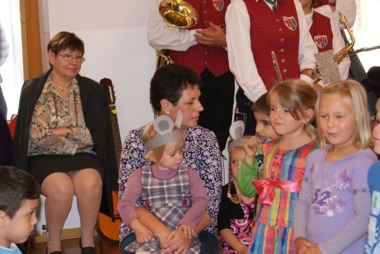 Eine Gruppe von Kindern und Erwachsenen versammelt sich drinnen, einige tragen Hasenkopfbänder. Eine Frau hält ein junges Mädchen, und ein Mann spielt im Hintergrund Saxophon.