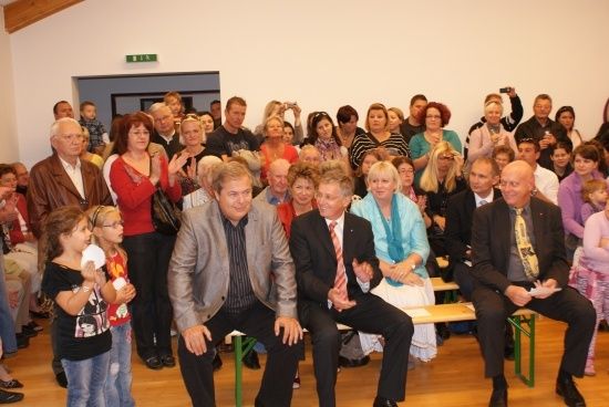 Eine Gruppe von Menschen, darunter mehrere Erwachsene und Kinder, sitzen und stehen in einem Raum, möglicherweise bei einer Veranstaltung. Einige klatschen, während andere Papiere halten. Ein Mann in Anzug und Krawatte sitzt auf einer Bank.