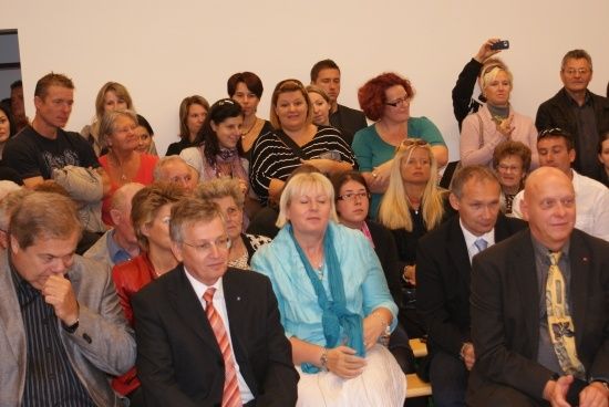 Eine Gruppe von Menschen ist in einem Raum versammelt, einige lächeln und andere schauen nach unten. Eine Frau in einem blauen Kleid sitzt in der Mitte.