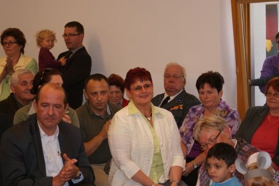 Eine Gruppe von Menschen ist in einem Raum versammelt, einige klatschen und andere schauen nach vorne. Eine Frau in der Mitte trägt eine Halskette und eine Uhr. Ein Kind sitzt auf dem Schoß von jemandem.
