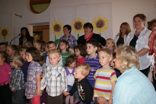 Eine Gruppe von Kindern und Erwachsenen steht in einem Raum mit einem Sonnenblumenbanner im Hintergrund. Die Kinder sind in verschiedenen Outfits gekleidet.