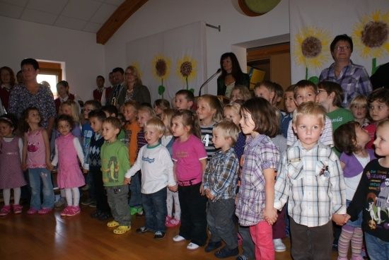 Eine Gruppe von Kindern und Erwachsenen steht in einem Raum mit Holzböden, Sonnenblumen-Bannern und einem Mikrofon auf einem Ständer.