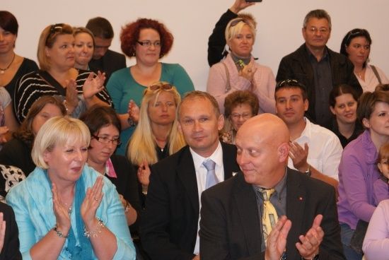 Eine Gruppe von Menschen, sowohl Männer als auch Frauen, klatschen und lächeln in einem Raum. Sie sind in formeller Kleidung gekleidet.
