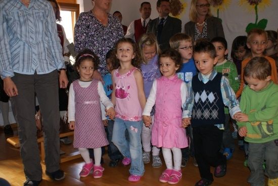 Eine Gruppe von Kindern in rosa Kleidern und Sandalen hält in einem Raum mit Erwachsenen Händchen.
