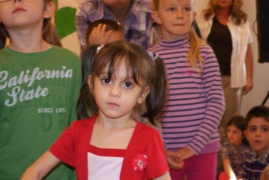 Eine Gruppe von Kindern steht in einem Raum, ein Mädchen in einem roten Shirt mit einer Blume in der Mitte schaut in die Kamera. Hinter ihr steht ein Mädchen in einem rosa gestreiften Shirt, das zur Seite schaut.