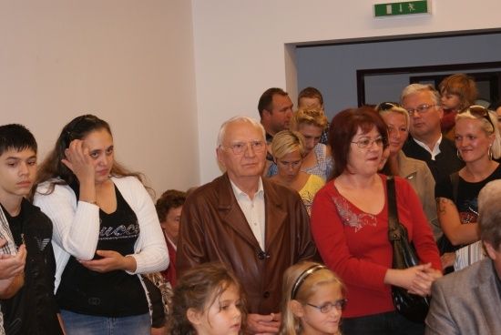 Eine Gruppe von Menschen, einschließlich Erwachsener und Kindern, steht in einem Raum, einige sehen ernst aus, andere lächeln. Ein Mann in einer braunen Jacke steht in der Mitte.