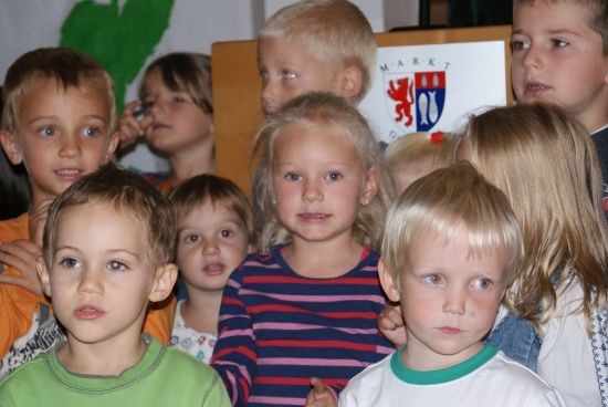 Eine Gruppe von Kindern, hauptsächlich Jungen, steht in einem Klassenzimmer zusammen. Sie schauen in verschiedene Richtungen. Ein Mädchen in der Mitte trägt ein gestreiftes Shirt. Hinter ihnen steht ein hölzerner Rednerpult mit einem Schild, auf dem 'MARKT' steht.