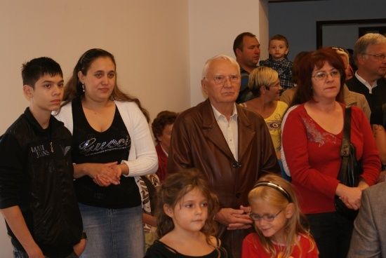 Eine Gruppe von Menschen, darunter ein Mann in einer braunen Jacke, steht in einem Raum zusammen. Eine Frau in einem schwarzen Shirt mit dem Wort Gucci darauf hält die Hand eines jungen Jungen.