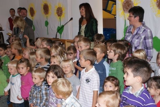 Eine Gruppe von Kindern und Erwachsenen versammelt sich in einem Raum, wobei eine Frau vor einem Mikrofon steht. Die Kinder schauen auf etwas vor sich. Der Raum hat eine weiße Wand mit gemalten Sonnenblumen.