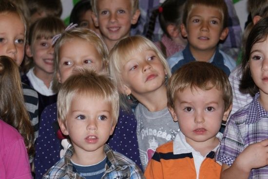 Eine Gruppe von Kindern, einige mit blonden Haaren, stehen zusammen und schauen in verschiedene Richtungen. Sie tragen verschiedenfarbige Shirts und scheinen sich in einem Klassenzimmer zu befinden.