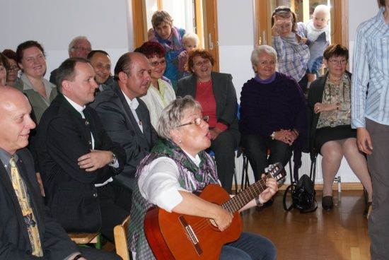 Eine Gruppe von Menschen versammelt sich in einem Raum, einige sitzen auf Stühlen und andere stehen. Eine Frau vorne spielt Gitarre, während andere zusehen.