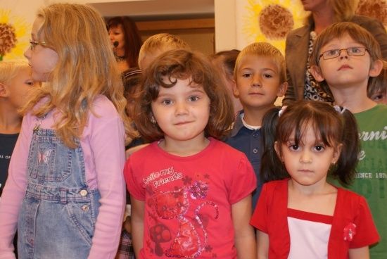 Eine Gruppe junger Kinder steht zusammen, einige mit Brille, mit einer Frau im Hintergrund. Das Mädchen in der Mitte trägt ein rotes Shirt mit Katzendesign.