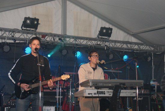 Zwei Männer spielen auf der Bühne, einer spielt Gitarre und der andere spielt ein Korg-Keyboard, unter blauen Bühnenlichtern.