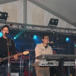 Zwei Männer spielen auf der Bühne, einer spielt Gitarre und der andere spielt ein Korg-Keyboard, unter blauen Bühnenlichtern.