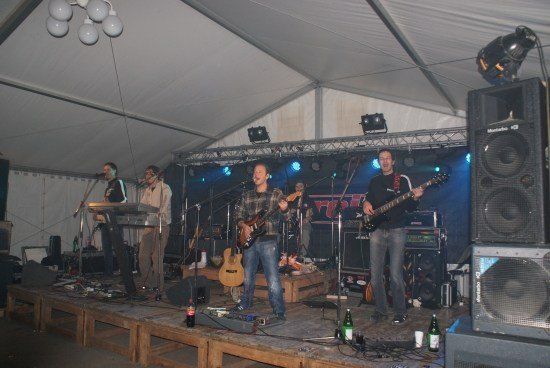 Eine Band spielt auf einer Bühne unter einem Zelt mit blauer Beleuchtung und Lautsprechern.