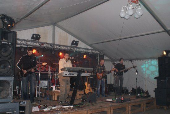 Eine Band spielt auf einer Bühne mit Zeltdach, bestehend aus einem Gitarristen, einem Keyboarder und zwei weiteren Gitarristen. Die Bühne ist mit Lautsprechern, Mikrofonen und Beleuchtung ausgestattet.
