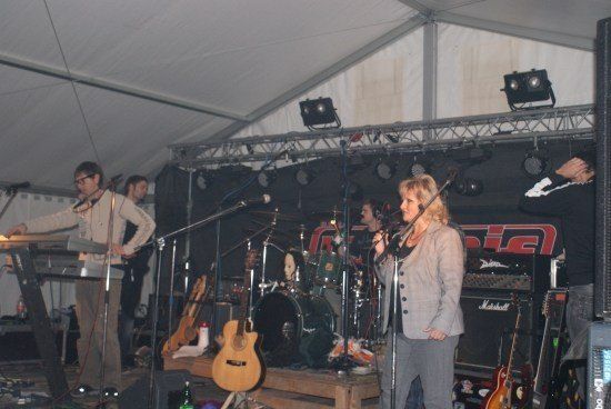 Eine Band spielt auf einer Bühne unter einem Zelt, mit einer Frau, die in ein Mikrofon singt, einem Mann, der das Keyboard spielt, und verschiedenen Gitarren und Trommeln aufgebaut.