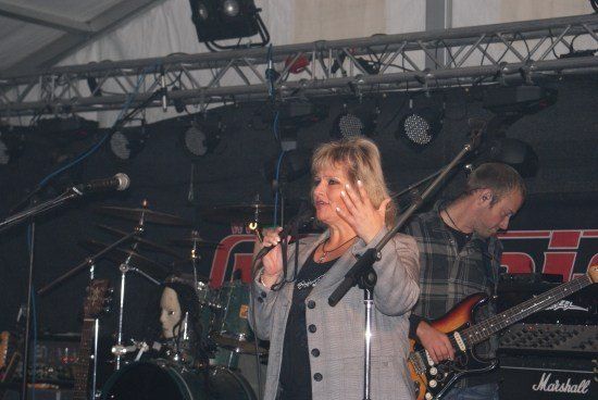 Eine Frau singt in ein Mikrofon auf der Bühne mit einer Band. Ein Mann spielt Gitarre hinter ihr.