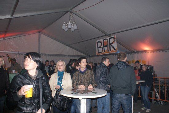 Eine Gruppe von Menschen versammelt sich in einem Zelt, einige stehen und einige sitzen. Eine Frau hält einen gelben Becher. Ein Schild oben zeigt 'BAR'.