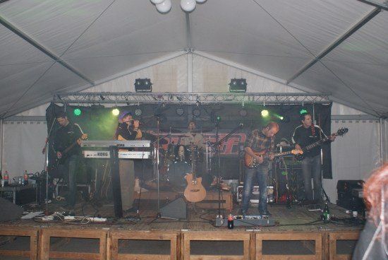 Eine Band spielt auf einer Bühne unter einem weißen Zelt mit grünen und weißen Lichtern. Die Band besteht aus einem Gitarristen, einem Keyboarder und einem Schlagzeuger.