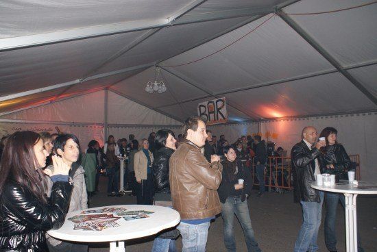 Eine Gruppe von Menschen versammelt sich in einem Zelt mit einem Bar-Schild. Einige stehen, andere sitzen an Tischen. Die Atmosphäre ist lebhaft mit Menschen, die reden und Gläser halten.