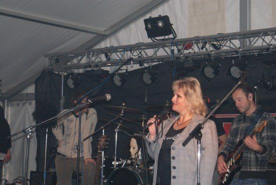 Eine Frau mit blonden Haaren singt in ein Mikrofon auf einer Bühne mit einer Band. Ein Mann spielt Gitarre hinter ihr.