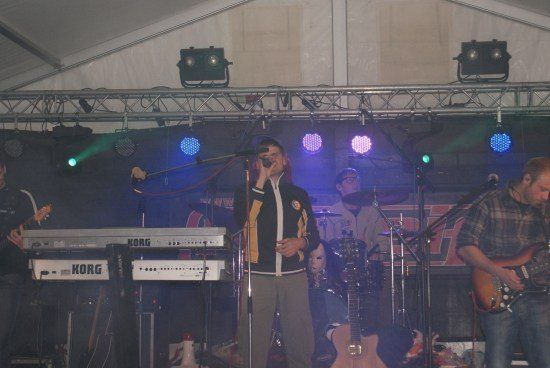 Eine Band spielt auf der Bühne mit einem Sänger, der in ein Mikrofon singt, einem Keyboarder, der ein Korg-Keyboard spielt, einem Gitarristen und einem Schlagzeuger. Die Bühne ist mit farbigen Lichtern beleuchtet und es gibt ein großes Banner hinter ihnen.