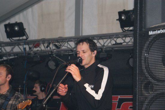 Ein Mann singt in ein Mikrofon auf der Bühne, mit einem Gitarristen und Schlagzeuger hinter ihm. Die Bühne ist mit Scheinwerfern beleuchtet und ein großer Lautsprecher ist sichtbar.