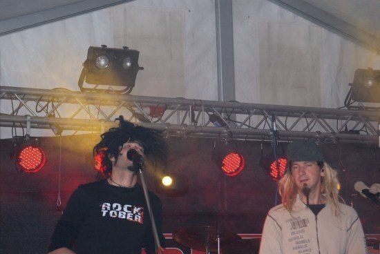 Zwei Männer treten auf der Bühne mit roten Lichtern und einem Mikrofon auf, einer trägt ein schwarzes Shirt mit weißem Text und der andere ein weißes Jackett.