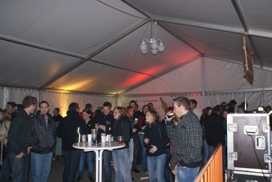 Eine Gruppe von Menschen ist in einem Zelt versammelt, einige trinken aus Tassen, während andere reden. In der Mitte steht ein Tisch mit Tassen und einer Flasche. Ein Mann hält eine Tasse und scheint zu trinken.