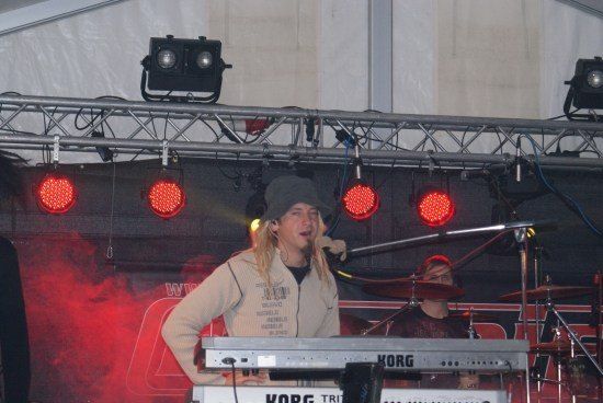 Ein Mann mit langen Haaren singt in ein Mikrofon auf einer Bühne mit roten Lichtern und einem Korg-Keyboard.