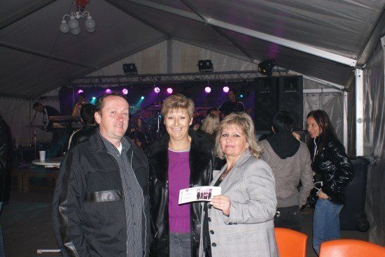 Drei Erwachsene stehen in einem Zeltbereich und lächeln für ein Foto. Eine Frau hält ein Ticket, und eine Band spielt auf der Bühne mit lila Beleuchtung.
