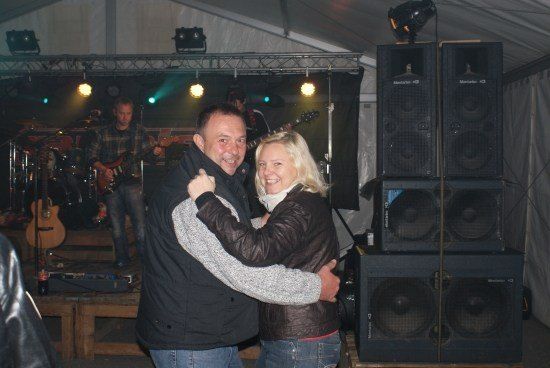 Ein Mann und eine Frau umarmen sich lächelnd vor einer Bühne, auf der eine Band auftritt. Die Bühne ist mit Lautsprechern, einem Schlagzeug und Gitarren ausgestattet.