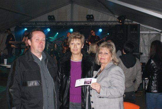 Drei Personen stehen vor einer Bühne mit einer Band in einem Zelt. Die Frau auf der rechten Seite hält ein Ticket.