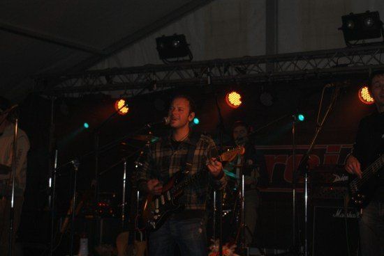 Eine Band spielt auf der Bühne unter Scheinwerfern, wobei der Gitarrist im Vordergrund eine E-Gitarre spielt und in ein Mikrofon singt.