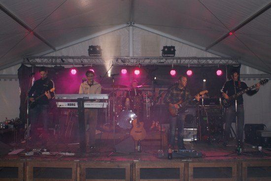 Eine Band spielt auf der Bühne unter einem Zelt mit rosa Beleuchtung. Sie spielen Gitarren, ein Keyboard und ein Schlagzeug.