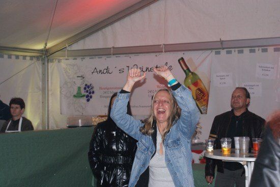 Eine Frau in einer Jeansjacke hebt ihre Hände in einem Zelt, möglicherweise bei einer Weinprobe. Hinter ihr steht ein Mann in einem schwarzen Jackett in der Nähe eines Tisches mit Getränken. Ein Banner mit dem Text 'Andi's 12ine' ist an der Wand.