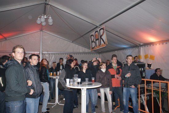 Eine Gruppe von Menschen versammelt sich in einem Zelt, möglicherweise auf einer Party. Ein Schild, das 'BAR' steht, hängt an der Decke. Einige Leute halten Getränke und Tassen. Ein Tisch mit Gläsern und Tassen steht in der Mitte. Eine Person steht in der Nähe des Tisches. Einige Leute stehen in der Nähe der Geländer. Eine Lampe hängt von der Decke.