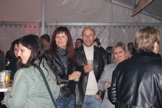 Eine Gruppe von Menschen steht in einem Raum, einige halten Getränke. Eine Frau in einer Lederjacke lächelt.