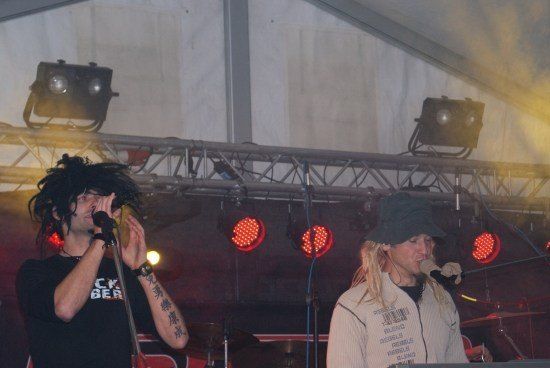 Zwei Personen stehen auf der Bühne und singen mit Mikrofonen, einer trägt ein schwarzes T-Shirt und der andere eine weiße Jacke. Sie stehen unter roten Bühnenlichtern und hinter ihnen steht ein Schlagzeug.