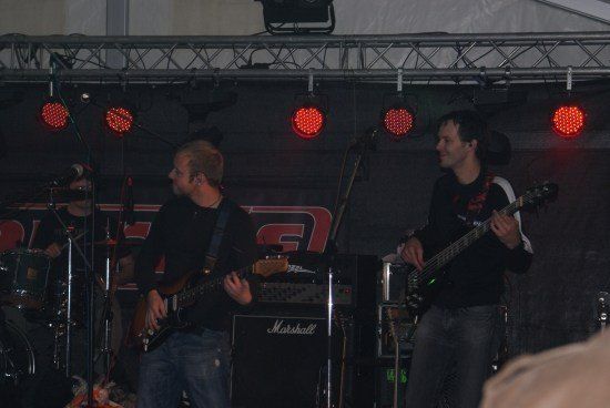 Eine Band spielt auf einer Bühne mit roter Beleuchtung, wobei ein Mann die Gitarre und ein anderer den Bass spielt.