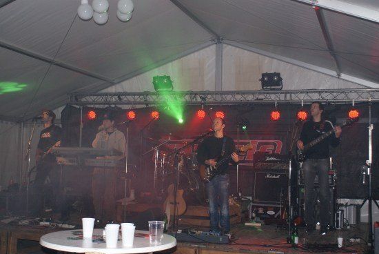 Eine Band tritt auf einer Bühne unter einem Zelt auf, das von grünen und roten Lichtern beleuchtet wird. Der Leadsänger singt in ein Mikrofon, während er die Gitarre spielt.